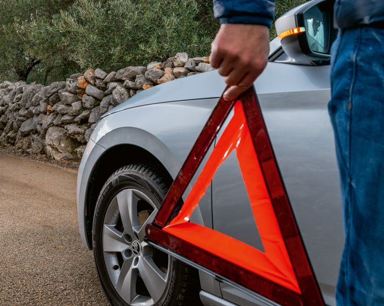 Ein oranger Škoda Karoq in der Hinteransicht auf einem Weg zwischen Olivenbäumen, im Vordergrund eine Hand, die ein Warndreieck hält.