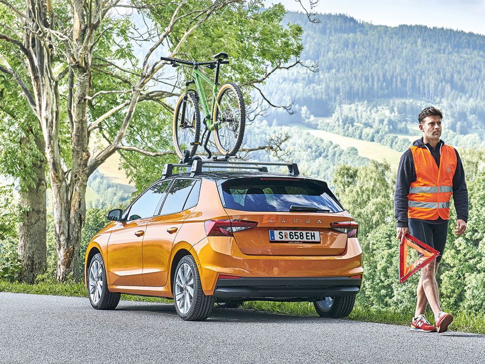 Ein Mann mit einer Warnweste und einem Pannendreieck marschiert von einen orangen Škoda fort.