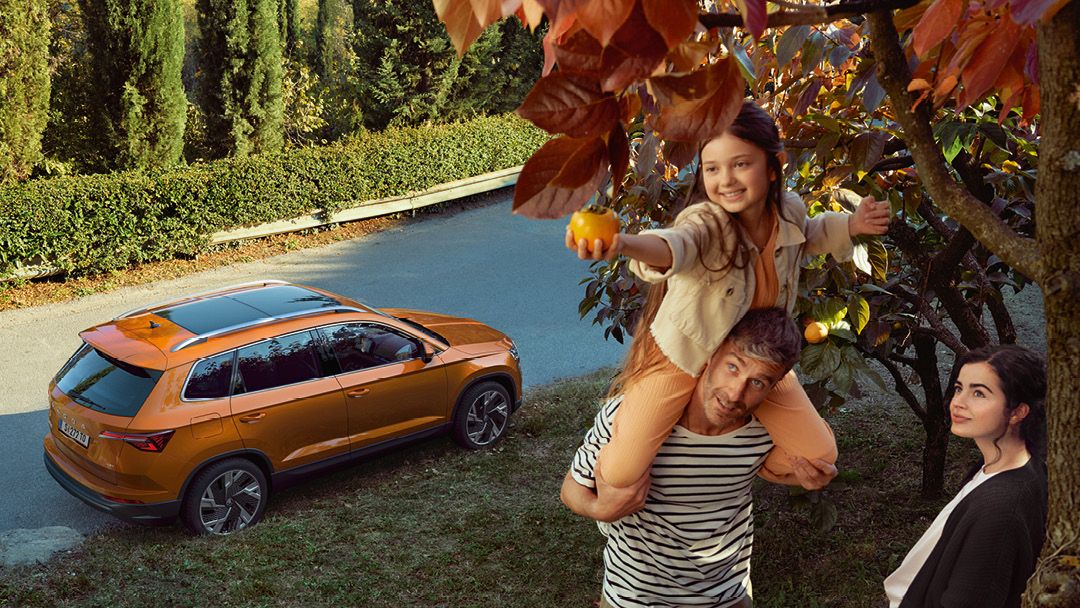 Eine Familie unter einem Baum, das Mädchen sitzt auf den Schultern des Vaters und pflückt Früchte. Im Hintergrund parkt ein Škoda Karoq.