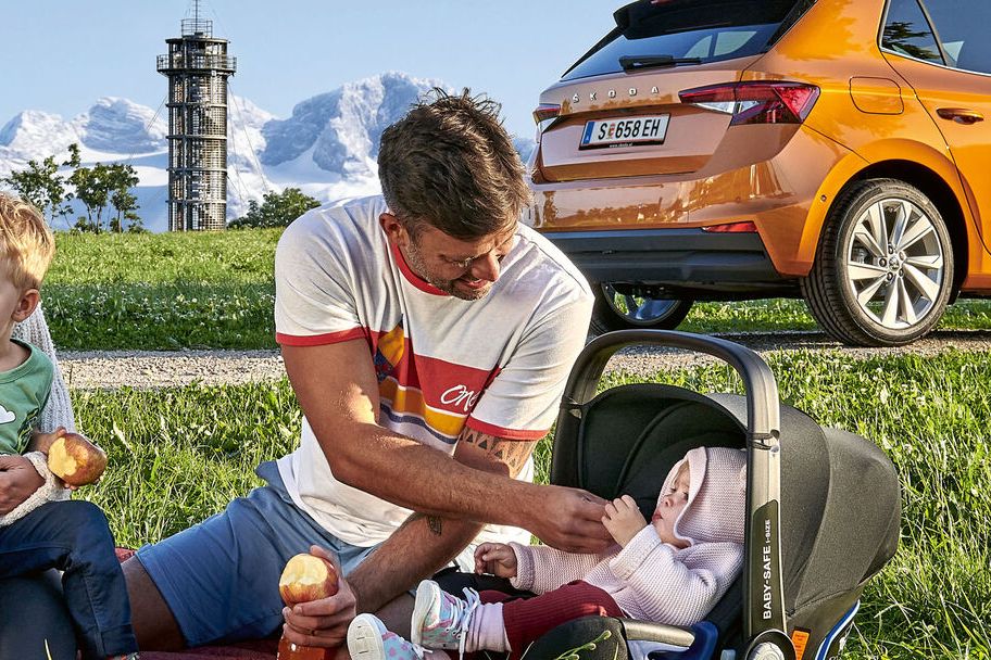 Eine Familie sitzt in der Landschaft vor einem orangen Skoda Fahrzeug.