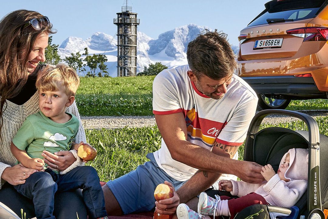 Eine Familie sitzt in der Landschaft vor einem orangen Skoda Fahrzeug.