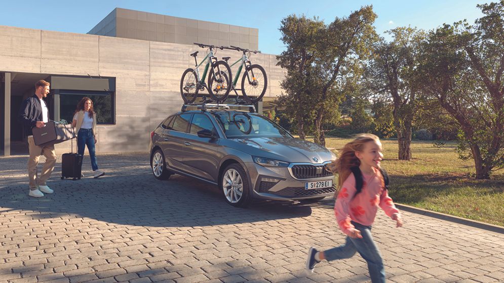 Ein grauer Škoda Scala mit Fahrraddachträger parkt vor einem Haus, die Eltern bereiten sich mit ihrer Tochter auf die Fahrt vor.