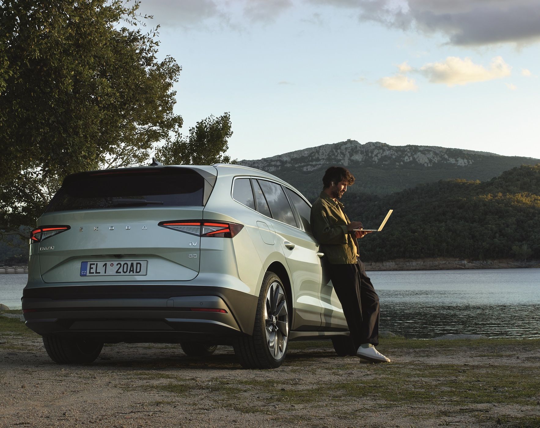 Ein Mann mit Laptop lehnt an einem Škoda Enyaq, dieser parkt an einem See